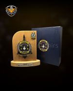 Wooden Commemorative Desk Plaque This wooden display plaque is built for official military and corporate presentations. The item features a solid light wood backboard mounted on a stable wooden base. It includes custom metal emblems and an engraving plate for specific event details. A matching presentation box accompanies the plaque for formal gifting. Product Specifications Build: Solid wood base and main board integrated with custom metal attachments. Detailing: Large central 3D metal crest and a secondary badge on the top right corner. Text Plate: Dedicated metal plate at the base for personalized text, names, and dates. Packaging: Secure presentation box featuring a custom logo on the lid to match the plaque design. Custom Production Details Wooden plaques with integrated metal badges are standard requirements for unit milestones, retirements, and corporate awards. The combination of custom wood shaping, detailed metal casting, and branded packaging highlights the manufacturing capacity available for specialized institutional orders. We produce these commemorative items according to exact unit and corporate specifications.